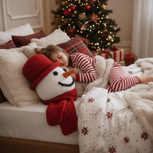 Op de afbeelding: Een kind slaapt in bed met een sneeuwman kussen. Het sneeuwman kussen heeft een rode hoed en sjaal, en een wortelneus. Het kind draagt een rood-wit gestreepte pyjama. Op de achtergrond is een kerstboom te zien.