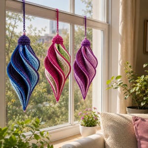 Könnte beinhalten: Drei spiralförmige Häkelornamente, die an einem Fenster hängen. Das erste ist blau und violett, das zweite pink und hellgrün, das dritte violett und hellrosa. Jedes Ornament hat eine gehäkelte Kappe und eine Aufhängekette.
