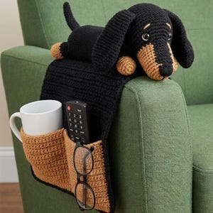 May include: A black and tan crochet dachshund dog armrest organizer with pockets for a mug, remote, and glasses. The organizer is draped over the arm of a green chair. The dachshund is lying on top of the armrest.