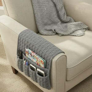 May include: A gray knitted armchair caddy with multiple pockets, holding a remote, pens, notebooks, and smartphones. The caddy is draped over the arm of a beige armchair, with a gray blanket draped over the back.