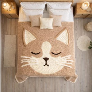 May include: A tan and cream-colored cat-face blanket on a bed. The blanket features a large, smiling cat face with closed eyes and whiskers. The bed is made with white sheets and several pillows. The room has wooden nightstands and flooring.