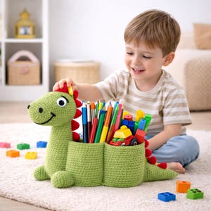 May include: A green, crocheted dinosaur desk tidy with red detailing, filled with pens, pencils, and small toys. The dinosaur has two compartments and a smiling face. Colourful blocks are scattered nearby.