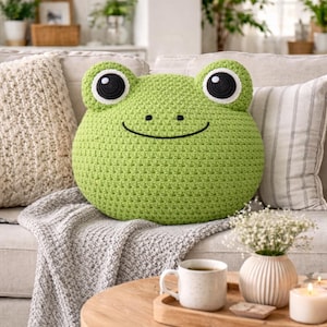 Könnte beinhalten: Ein großes, rundes, grünes gehäkeltes Froschkissen mit schwarz-weißen Augen und einem lächelnden Mund. Das Froschkissen liegt auf einer grauen Decke auf einem beigen Sofa, mit anderen Kissen im Hintergrund. Dekorative Wohnkultur.