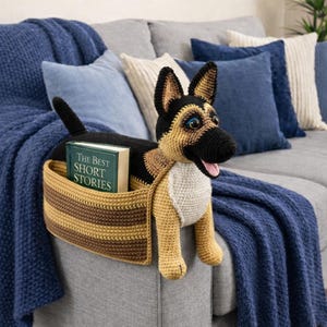 Puede incluir: Organizador de brazo de sofá con forma de perro pastor alemán tejido a ganchillo en negro, marrón y crema. El perro tiene un bolsillo para libros con un libro titulado "The Best Short Stories". El perro está sentado en un sofá gris con cojines azules y una manta azul.