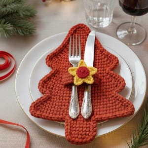 Puede incluir: Una mesa navideña festiva con un salvamanteles en forma de árbol de ganchillo de color óxido. La cubertería, que incluye un tenedor y un cuchillo, se sujeta con una flor de ganchillo amarilla y roja. El conjunto está sobre un plato blanco.