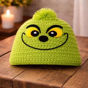 Peut inclure: Un bonnet vert crochet&eacute; avec un motif de visage de dessin anim&eacute;. Le chapeau pr&eacute;sente des yeux jaunes, un nez noir et une bouche souriante. Un pompon vert est sur le dessus. Le chapeau est pos&eacute; sur une surface en bois.
