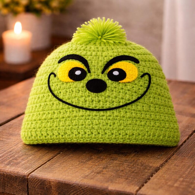 Peut inclure: Un bonnet vert crochet&eacute; avec un motif de visage de dessin anim&eacute;. Le chapeau pr&eacute;sente des yeux jaunes, un nez noir et une bouche souriante. Un pompon vert est sur le dessus. Le chapeau est pos&eacute; sur une surface en bois.