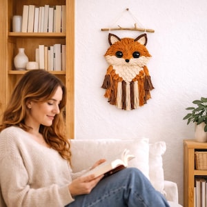 May include: A handcrafted wall hanging featuring a fox design. The fox is made of yarn in shades of orange, brown, and cream, with large black eyes. It is suspended from a wooden branch with a white string, adding a rustic touch to the decor.