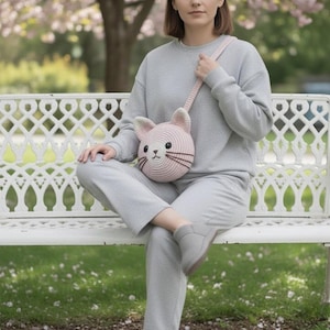 Puede incluir: Un bolso bandolera de ganchillo rosa claro con forma de gato y una correa a juego. El bolso presenta bigotes y ojos negros, y orejas internas de color rosa claro. El bolso se lleva al hombro.