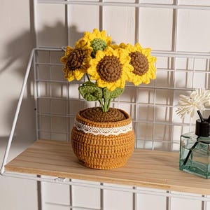 May include: A crochet sunflower arrangement featuring three bright yellow flowers with dark brown centers and green leaves, potted in a textured mustard-colored yarn container adorned with a white lace trim around the upper edge.