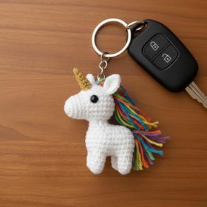 May include: A white crochet unicorn keychain with a gold horn and a rainbow mane. The keychain is attached to a silver ring, next to a black car key on a wooden surface. The unicorn has black eyes.