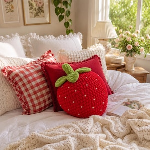 May include: A plush, strawberry-shaped throw pillow in bright red with a textured, yarn-like surface, small white pearl-like accents, and a green leafy top. It is positioned on a bed alongside a red and white gingham pillow and a white blanket.