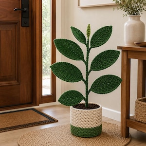 Pode incluir: Uma planta de crochê com um caule central, várias folhas verde-escuras e um pequeno broto verde-claro no topo. A planta está num vaso cilíndrico com um padrão texturizado de estilo trançado em fios de cor creme e verde-escura.