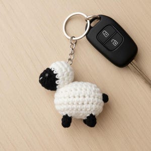 May include: A handmade crochet sheep keychain with a silver keyring. The sheep is white with a black head, legs, and tail. A black car key is next to the keychain.