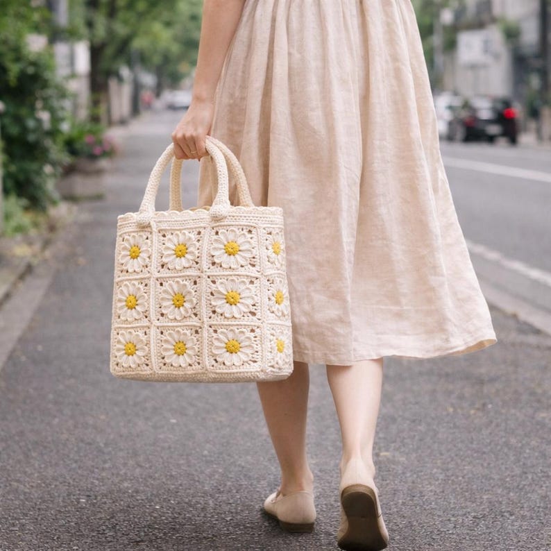 Op de afbeelding: Cr&egrave;mekleurige gehaakte tote bag met een madeliefjespatroon. De tas heeft een vierkant ontwerp met gele en witte madeliefjesmotieven. De handvatten zijn ook gehaakt. De tas wordt vastgehouden door een persoon die op een verharde weg loopt.