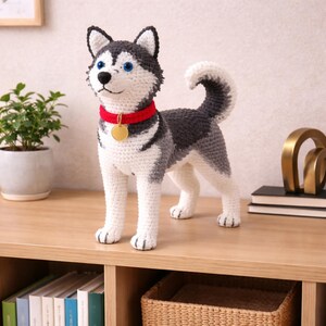 May include: A handmade crochet Husky dog figurine. The dog has a red collar with a gold tag, blue eyes, and a black nose. The dog's fur is white, gray, and black. The figurine is standing on a wooden surface.