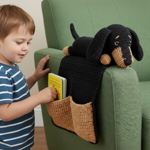 May include: A black and tan crochet dachshund dog armrest organizer with two pockets. The dog is lying on top of a black fabric that hangs over the armrest. A child is reaching into one of the pockets, which contains a small book.
