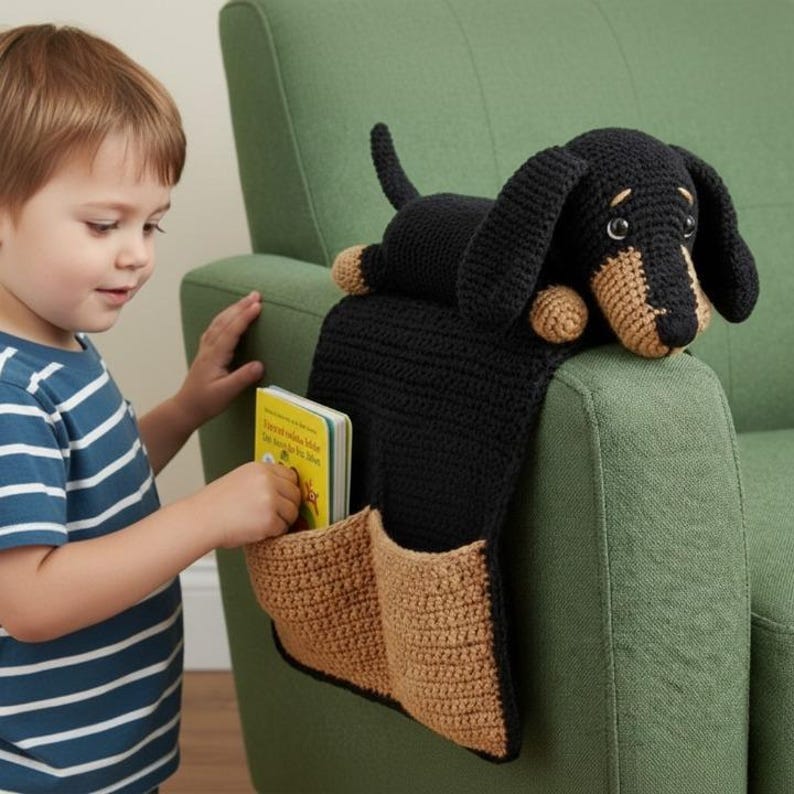 May include: A black and tan crochet dachshund dog armrest organizer with two pockets. The dog is lying on top of a black fabric that hangs over the armrest. A child is reaching into one of the pockets, which contains a small book.