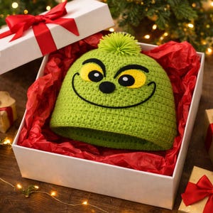Peut inclure: Un bonnet vert en crochet avec un motif de visage de dessin anim&eacute;, avec des yeux jaunes, un nez noir et une bouche souriante. Le chapeau est pr&eacute;sent&eacute; dans un coffret cadeau blanc avec du papier de soie rouge et un n&oelig;ud rouge.