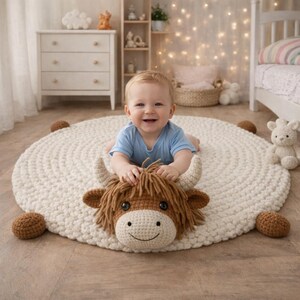 May include: A smiling baby in a blue onesie lies on a cream-colored, round, crocheted rug with a brown and tan Highland cow head. The rug has four brown crocheted balls around the edge. A white dresser and bed are in the background.