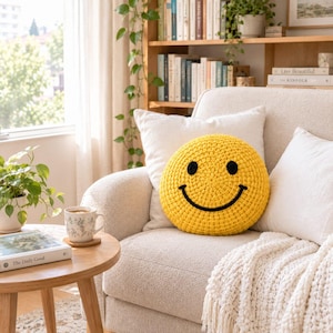 May include: A round, bright yellow crochet pillow featuring a black stitched smiley face with two circular eyes and a curved mouth. The pillow is textured with visible yarn loops and is positioned on a light-colored sofa with white cushions.