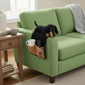 May include: A black and tan dachshund-shaped armrest organizer with a cup holder, remote control pocket, and glasses holder. The organizer is placed on the arm of a green sofa. A matching dachshund plush toy rests on top.