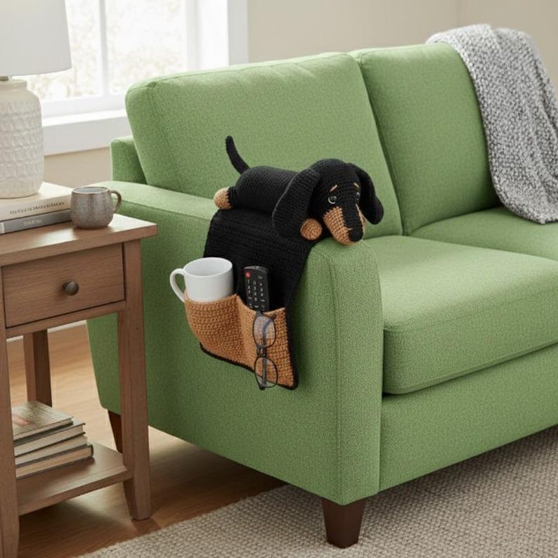 May include: A black and tan dachshund-shaped armrest organizer with a cup holder, remote control pocket, and glasses holder. The organizer is placed on the arm of a green sofa. A matching dachshund plush toy rests on top.