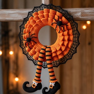Puede incluir: Una corona de Halloween tejida a ganchillo con un centro naranja con volantes, un borde negro similar a una telaraña y dos arañas negras. Dos piernas largas y colgantes con rayas horizontales naranjas y negras terminan en zapatos negros de punta curva.