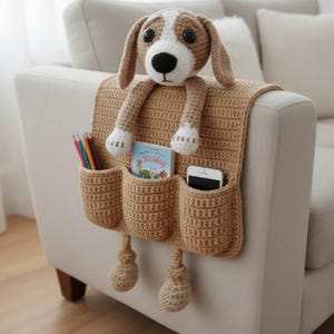 May include: A tan crochet couch tidy with three pockets and a stuffed dog head and legs. The organiser is attached to the arm of a beige sofa. Pencils, a book, and a smartphone are in the pockets.