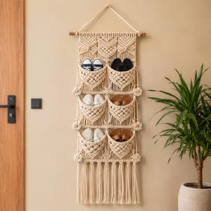 Può includere: Un organizer da parete in macramè color crema con sei tasche individuali disposte su due colonne. L'organizer è appeso a un bastone di legno e presenta decorazioni floreali annodate e lunghe frange nella parte inferiore.