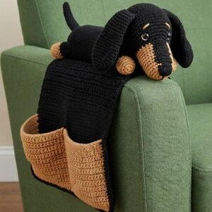 May include: A black and tan crochet dachshund dog armrest organizer with two pockets. The dog is lying on top of the armrest, with its head and paws visible. The pockets are tan and the rest of the organizer is black. The organizer is on a green chair.