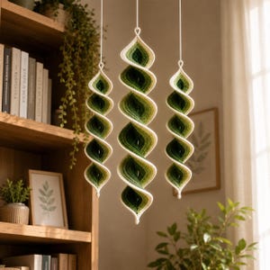 May include: Three hanging spiral ornaments crafted from yarn in an ombre gradient of forest green, sage green, and cream. Each piece features a twisted, elongated shape suspended by a thin white cord, creating a vertical display of color.
