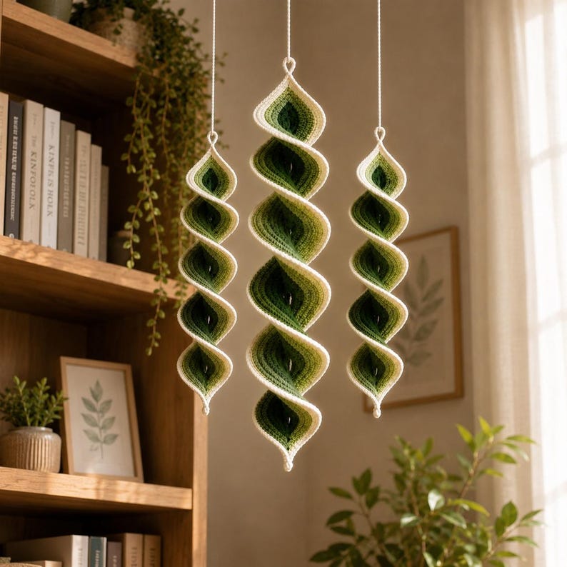 May include: Three hanging spiral ornaments crafted from yarn in an ombre gradient of forest green, sage green, and cream. Each piece features a twisted, elongated shape suspended by a thin white cord, creating a vertical display of color.