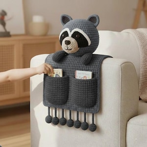 May include: A grey crocheted sofa arm organiser with a raccoon head and two pockets. The raccoon has a black mask, white accents, and small ears. The organiser has two pockets and decorative pom-poms hanging from the bottom. A child's hand is reaching into a pocket.