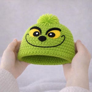 Peut inclure: Un bonnet vert crochet&eacute; avec un visage de dessin anim&eacute;, comprenant des yeux jaunes, un nez noir et une bouche souriante. Le chapeau est surmont&eacute; d'un pompon vert. Le bonnet est tenu &agrave; deux mains.
