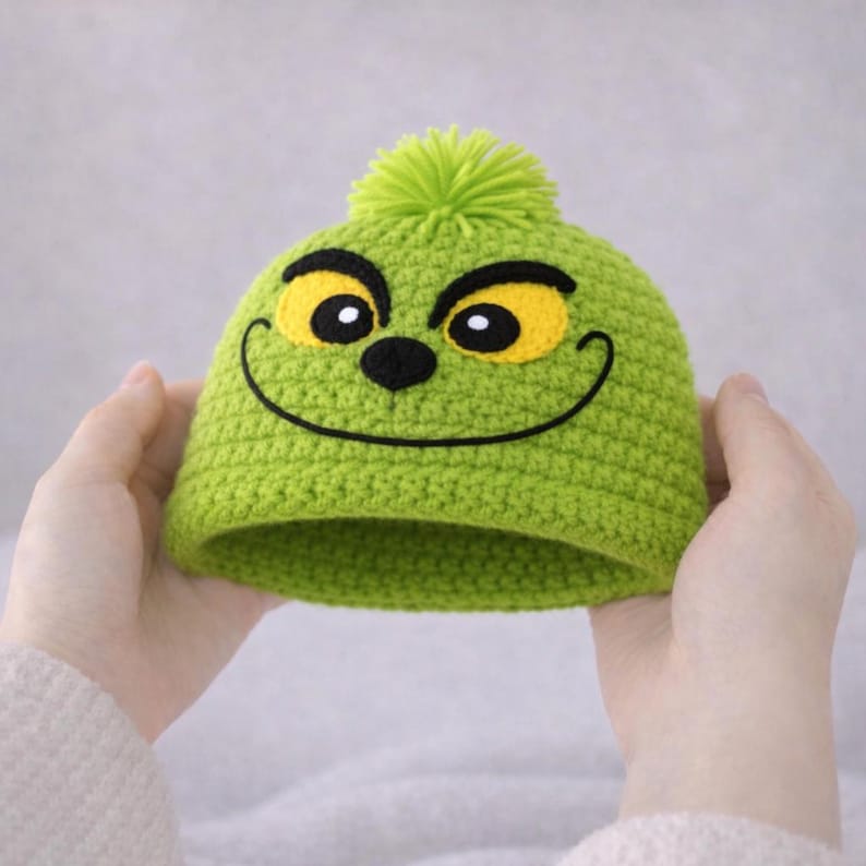 Peut inclure: Un bonnet vert crochet&eacute; avec un visage de dessin anim&eacute;, comprenant des yeux jaunes, un nez noir et une bouche souriante. Le chapeau est surmont&eacute; d'un pompon vert. Le bonnet est tenu &agrave; deux mains.