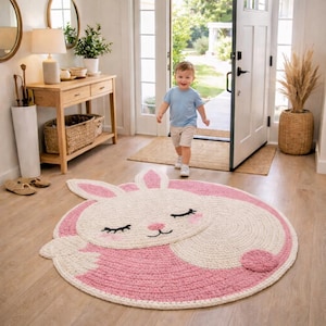 May include: A round, pink and white bunny rug with a smiling face and closed eyes. The rug is made of a thick, woven material and is placed on a wooden floor. The rug is a decorative item for a child's room.
