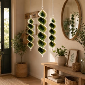May include: Three hanging spiral ornaments crafted from yarn in an ombre gradient of forest green, olive, and cream. Each piece features a twisted, conical shape suspended by a thin white string, showcasing a textured, hand-crocheted appearance.