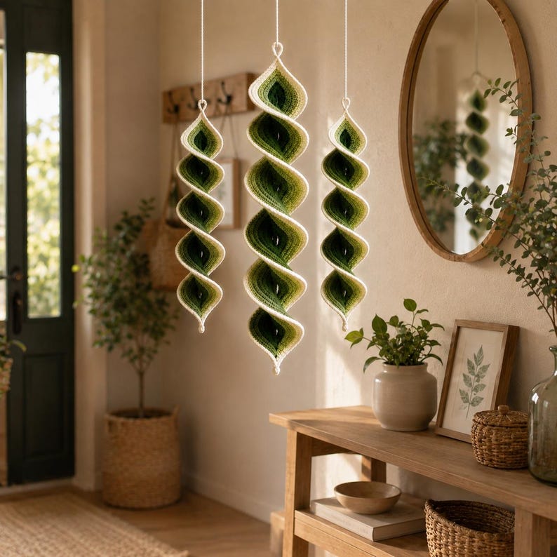 May include: Three hanging spiral ornaments crafted from yarn in an ombre gradient of forest green, olive, and cream. Each piece features a twisted, conical shape suspended by a thin white string, showcasing a textured, hand-crocheted appearance.