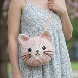 Puede incluir: Un bolso bandolera rosa tejido a ganchillo con forma de gato, con una correa larga. El bolso presenta una cara de gato con ojos negros, bigotes marrones y una nariz peque&ntilde;a. Las orejas son de un rosa m&aacute;s claro con puntas blancas. El bolso se lleva al hombro.