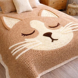 May include: A tan and cream-colored cat-shaped bedspread. The bedspread features a large cat face with closed eyes, whiskers, and a nose. The bedspread is made of a textured material and is placed on a bed with matching pillows.