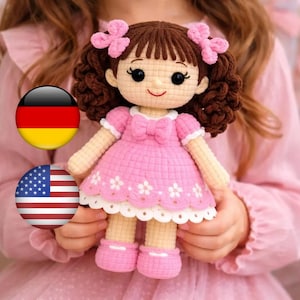 May include: A handmade crochet doll with brown curly hair, pink bows, and a pink dress with white floral accents. The doll has black eyes and is wearing pink shoes. The doll is being held by a person.