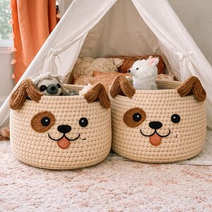 May include: Two beige crochet dog-shaped storage baskets with brown ears, eyes, and spots. Each basket has a smiling face and a pink tongue. One basket contains a stuffed sloth, and the other contains a stuffed white dog.