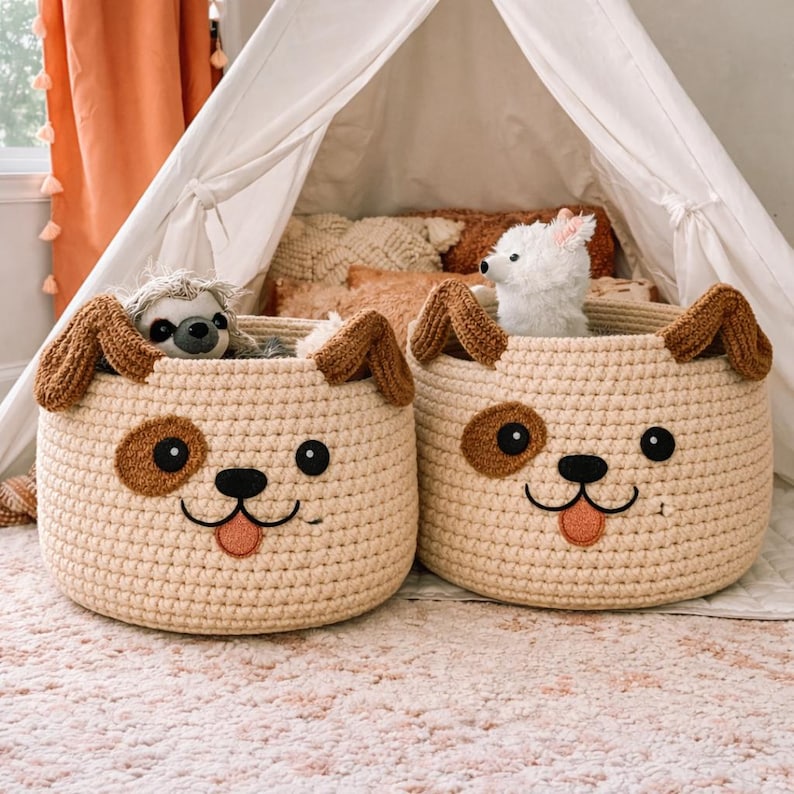May include: Two beige crochet dog-shaped storage baskets with brown ears, eyes, and spots. Each basket has a smiling face and a pink tongue. One basket contains a stuffed sloth, and the other contains a stuffed white dog.