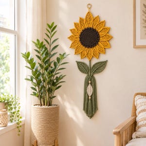 Könnte beinhalten: Ein Makramee-Wandbehang in Form einer Sonnenblume mit gelben Blütenblättern, einem dunkelbraunen Zentrum und einem grünen Stiel mit zwei Blättern sowie einem hängenden cremeweißen Blattelement. Das Objekt ist an einem kleinen goldenen Ring befestigt.