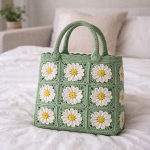 Op de afbeelding: Een groene gehaakte tote bag met witte madeliefjes applicaties en gele centra. De tas heeft twee afgeronde handvatten en is gemaakt van vierkante gehaakte panelen. De tas staat op een witte ondergrond.