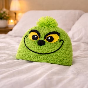 Peut inclure: Un bonnet vert crochet&eacute; avec un motif de visage de dessin anim&eacute;. Le chapeau pr&eacute;sente des yeux jaunes avec des contours noirs, un nez noir et un sourire noir cousu. Un pompon vert est plac&eacute; au sommet du chapeau.