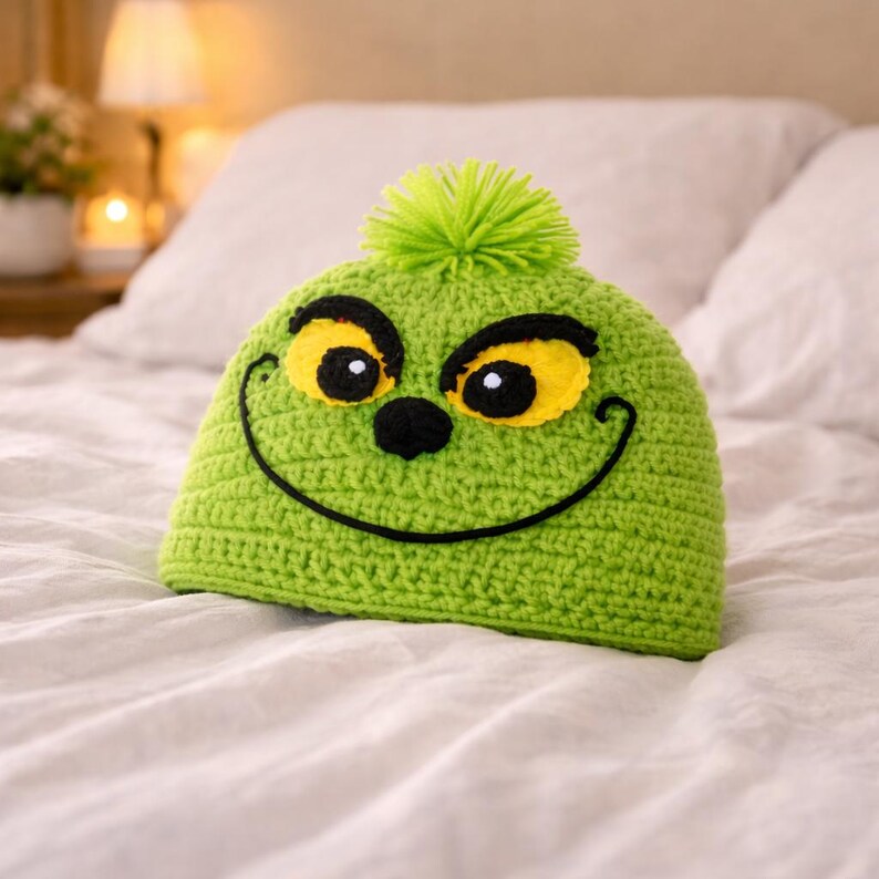 Peut inclure: Un bonnet vert crochet&eacute; avec un motif de visage de dessin anim&eacute;. Le chapeau pr&eacute;sente des yeux jaunes avec des contours noirs, un nez noir et un sourire noir cousu. Un pompon vert est plac&eacute; au sommet du chapeau.