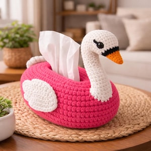 May include: A handmade crochet tissue box cover shaped like a swan. The swan has a bright pink body, white wings, and a white head with a black and orange beak. White tissues are visible coming out of the swan's body.