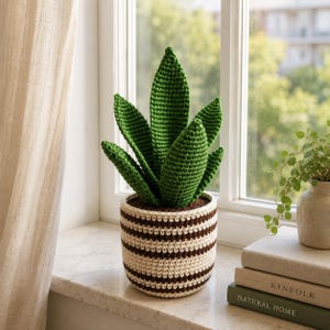 May include: A crocheted snake plant with upright, pointed green leaves arranged in a cluster. The plant is housed in a cylindrical, crocheted pot featuring horizontal stripes in alternating cream and dark brown yarn colors.